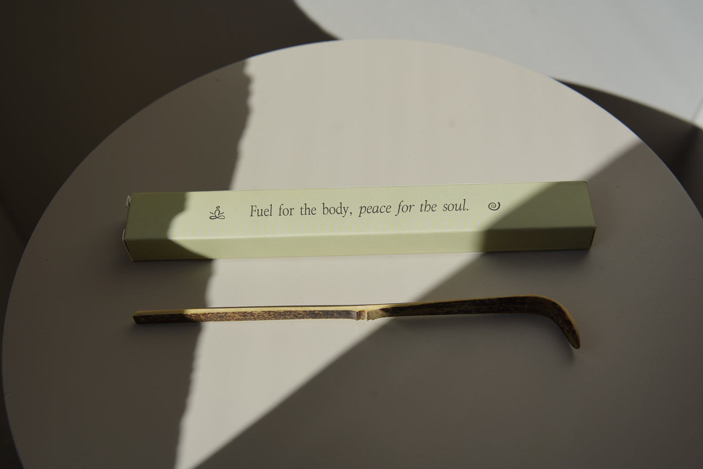 Matcha bamboo scoop and green box with the text “Fuel for the body, peace for the soul” placed on a round white table in sunlight.