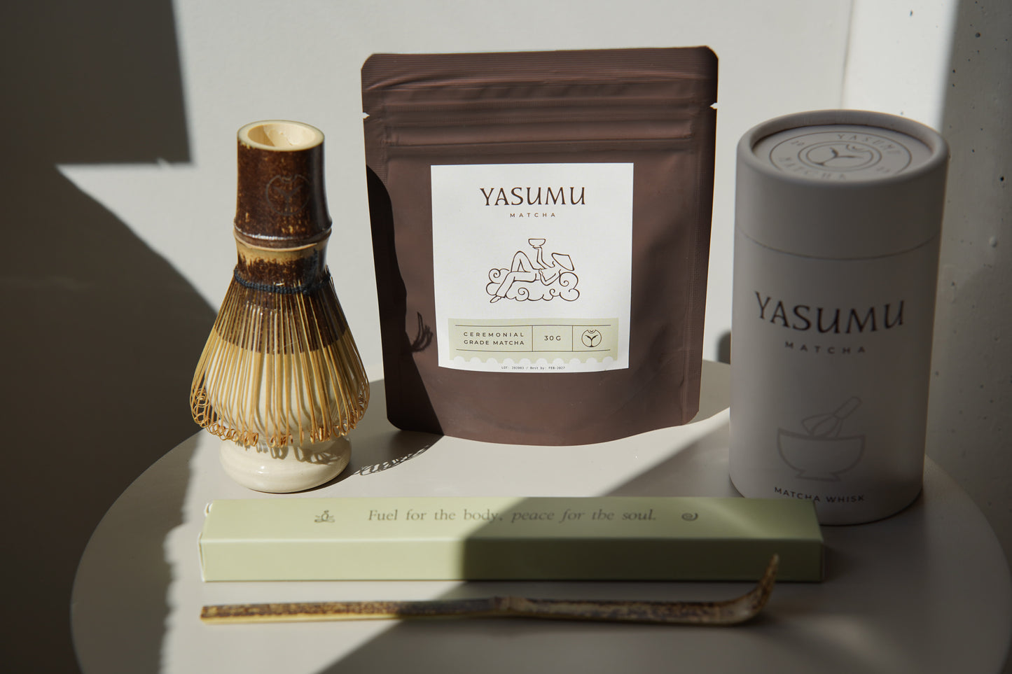 Yasumu Matcha complete set with bamboo whisk, ceremonial matcha pouch, white matcha tin, and bamboo scoop box displayed on round white table in natural light