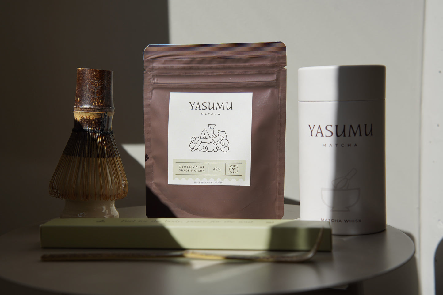 Yasumu Matcha complete set with bamboo whisk, ceremonial matcha pouch, white matcha tin, and bamboo scoop box displayed on round white table in natural light