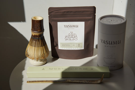 Yasumu Matcha complete set with bamboo whisk, ceremonial matcha pouch, white matcha tin, and bamboo scoop box displayed on round white table in natural light