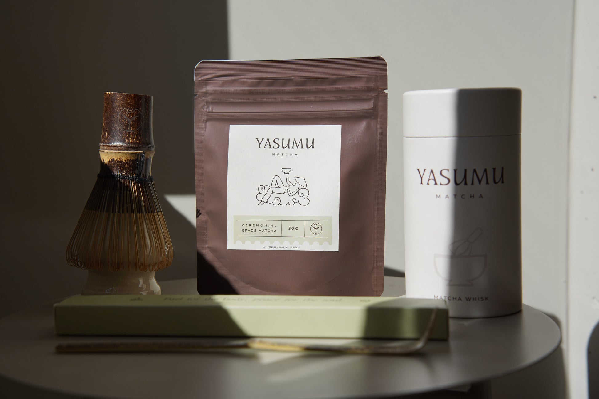 Yasumu Matcha complete set with bamboo whisk, ceremonial matcha pouch, white matcha tin, and bamboo scoop box displayed on round white table in natural light