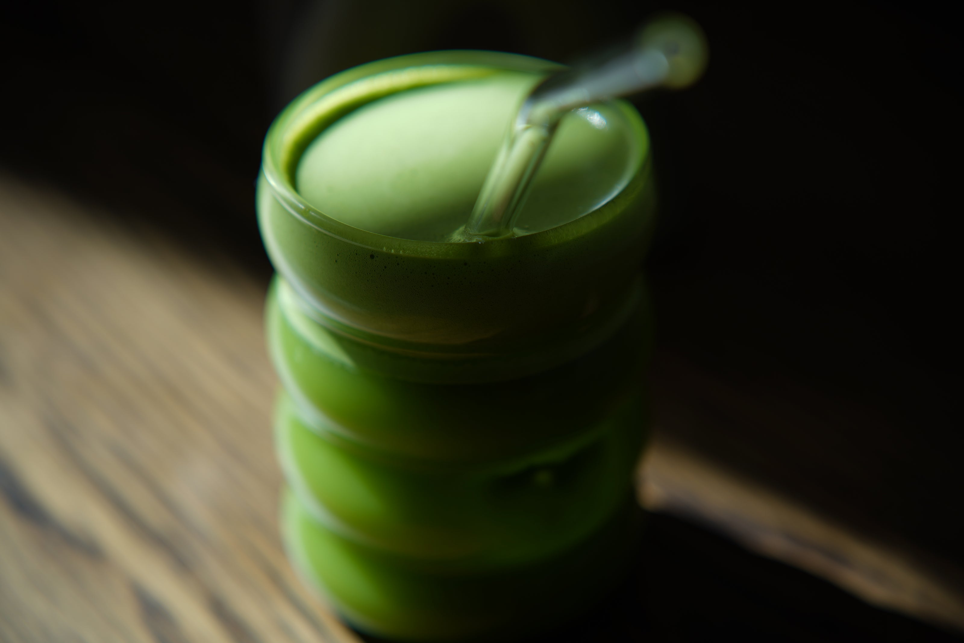 Close-up of creamy ceremonial matcha latte in bright green wavy glass with glass straw on wooden surface