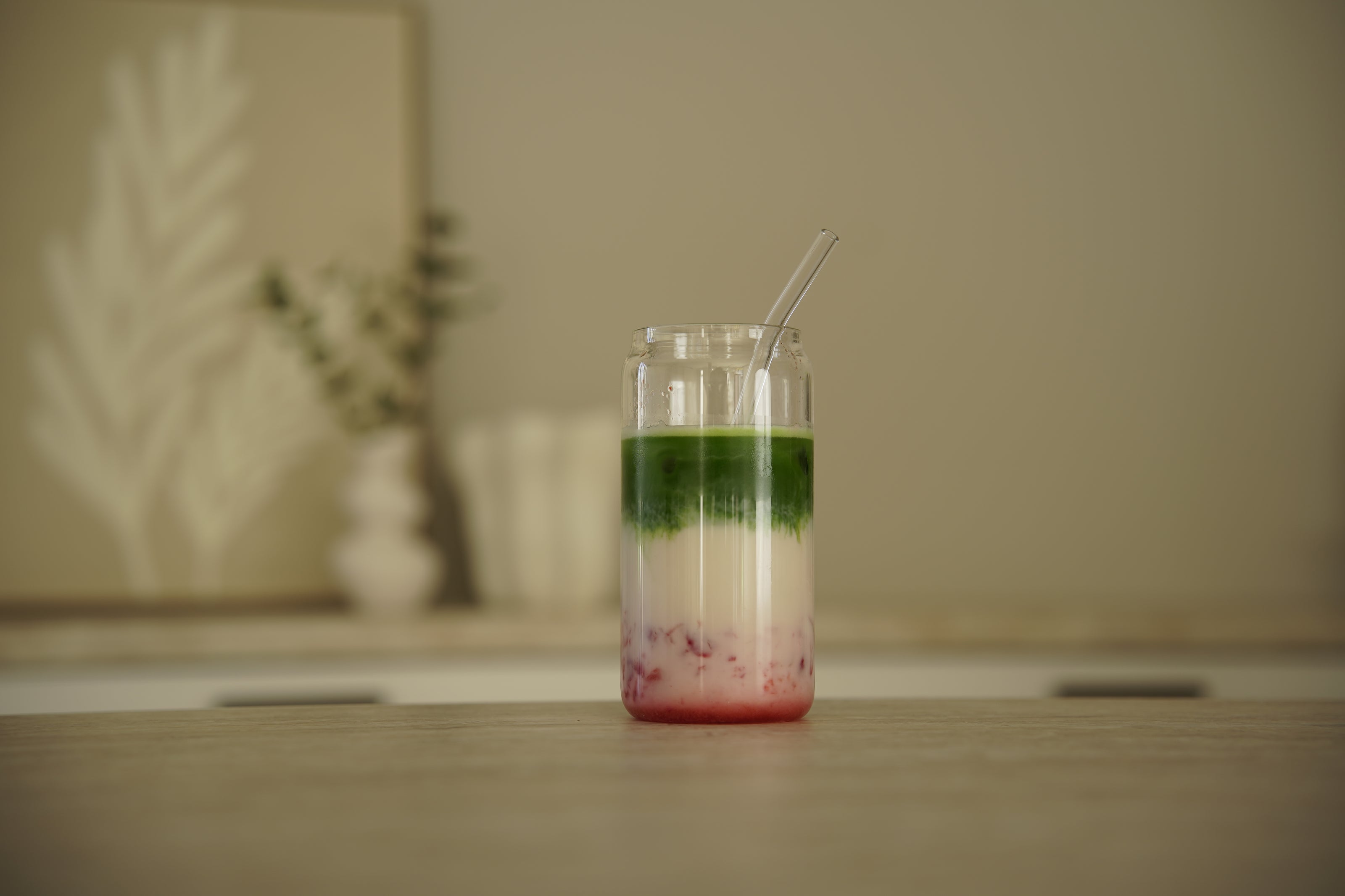 Layered matcha strawberry latte in clear glass with glass straw on light wooden table in minimalist kitchen setting