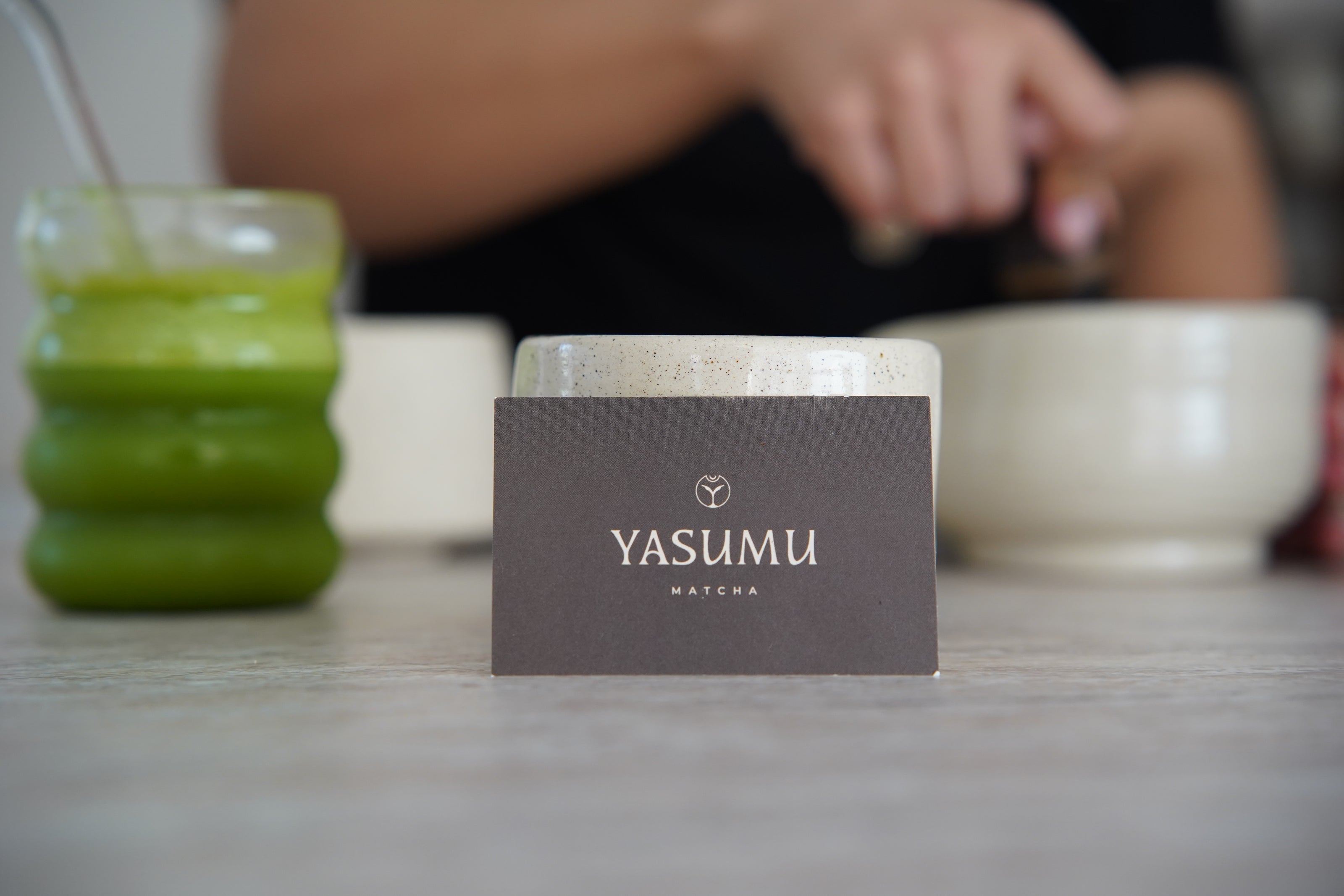 Yasumu Matcha branded card on table with iced matcha latte and person whisking matcha in ceramic bowls in background