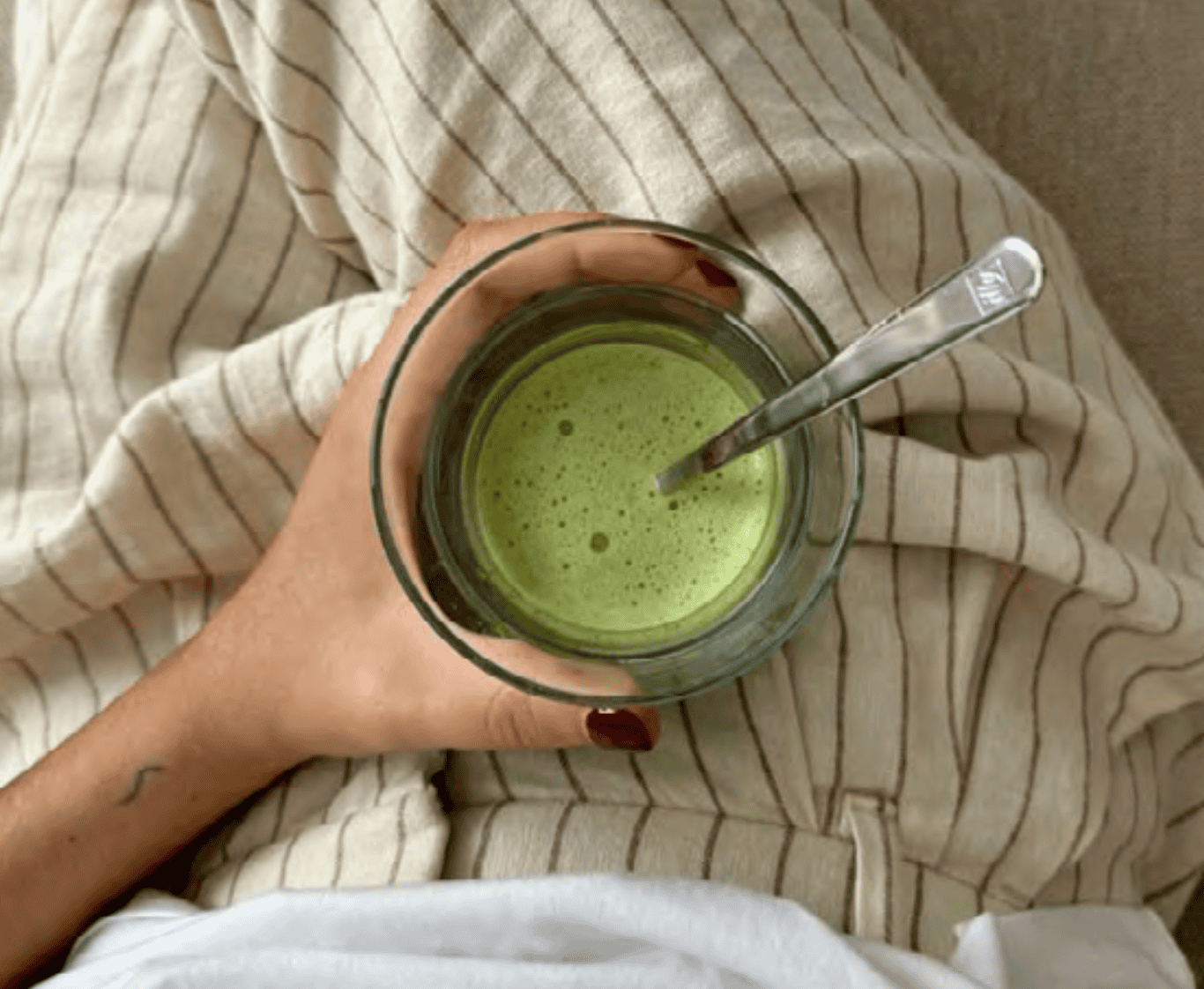 Top view of a hand holding a glass of freshly whisked matcha with a spoon, resting on striped beige loungewear.
